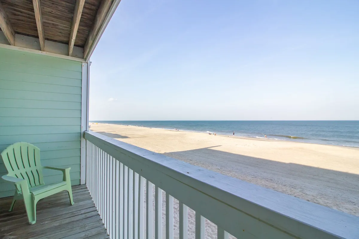 A serene view of the beach and ocean from a balcony, featuring a green chair inviting guests to relax. The setting is ideal for sipping a drink and soaking in sunset or sunrise views, blending private comfort with natural beauty.