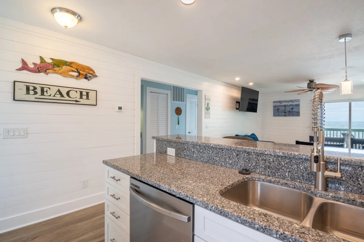This image features a modern kitchen with a granite countertop, beach-themed decor, and contemporary appliances. The open layout and visible beach sign infuse a sense of coastal charm, while the design accommodates entertaining friends or relaxing with family.