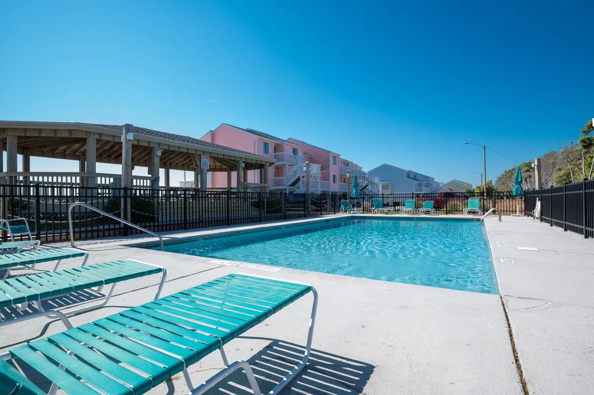 A spacious outdoor swimming pool surrounded by inviting lounge chairs and bordered by the community's resort-style buildings. The area is well-maintained, offering plenty of space for sunbathing, swimming, and relaxing with family or friends under blue skies.
