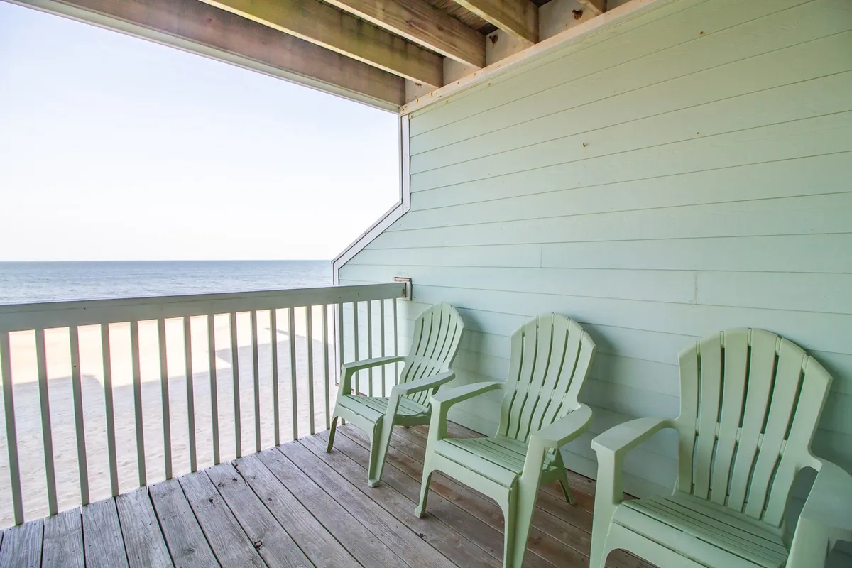 An exterior shot featuring green Adirondack chairs on a wooden deck facing the ocean. The view is open and expansive, offering a clear line of sight to the water and the horizon. The setting is perfect for sipping drinks, birdwatching, or quietly soaking in the sunrise and ocean breezes from the property itself.