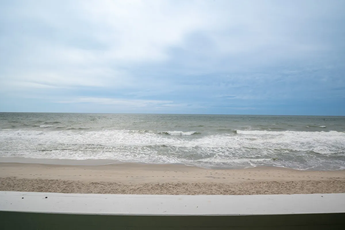 A wide, unobstructed view of the sandy beach with gentle ocean waves rolling in under a cloudy sky. The shoreline is open and perfect for family activities like beach walks, collecting seashells, and building sandcastles.
