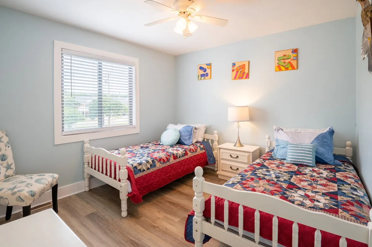 A cheerful second bedroom with twin beds adorned in colorful quilts, a nightstand with lamp, and a window with blinds. The space feels cozy and inviting, ideal for kids or friends to relax, read, or play, with plenty of natural light enhancing the comfort.