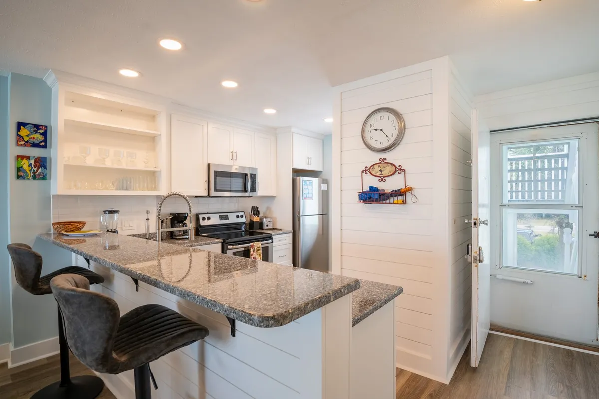 This image presents a remodeled kitchen featuring a granite countertop, stainless steel appliances, bar stools for three, and bright, cheerful wall art. The open design invites interaction between the kitchen and adjoining spaces, and the bar seating is conveniently positioned for casual dining or conversation.