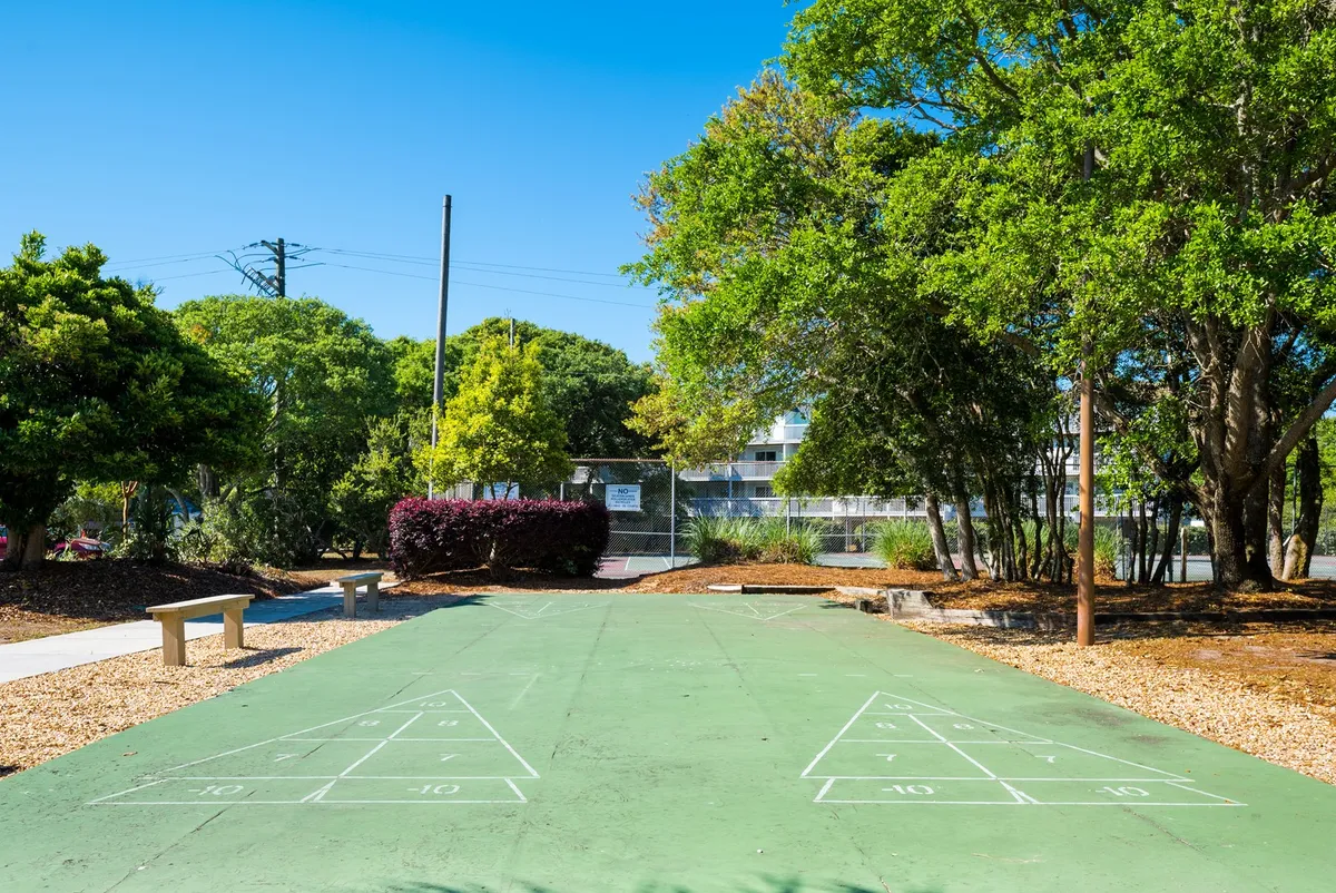 This image features an exterior view of a shuffleboard court, framed by lush greenery and benches where guests can relax. The setting is casual and playful, inviting friendly competitions and social gatherings in a pleasant outdoor environment.