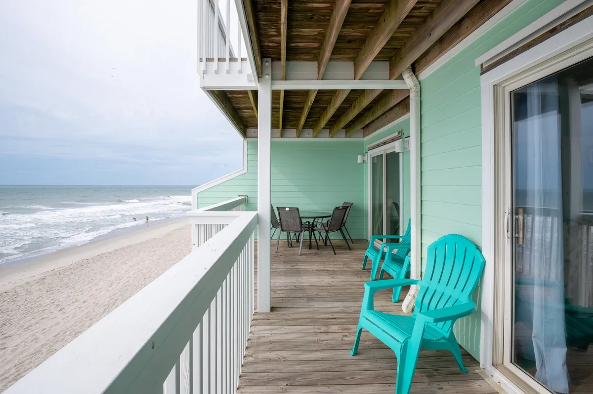 An exterior deck scene with turquoise chairs, a wooden deck, and a patio table, all overlooking a beautiful beachfront view. The scene invites relaxation, morning coffee, or sunset viewing, with the endless ocean forming a stunning backdrop.