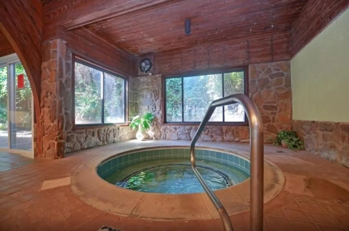 A serene interior hot tub area with stone walls, lush indoor plants, and large windows letting in natural light. The atmosphere is tranquil and spa-like, perfect for relaxation during rainy days or the off-season.