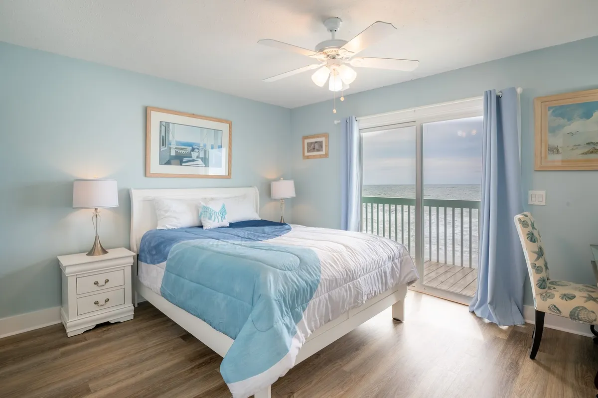 The image shows a serene bedroom with soft blue and white bedding, a large window framing sweeping ocean views, a cozy nightstand with a lamp, and the promise of tranquil mornings. The sunlit space feels airy and calm, inviting guests to unwind, read by the window, or savor a peaceful coffee while gazing at the sea.