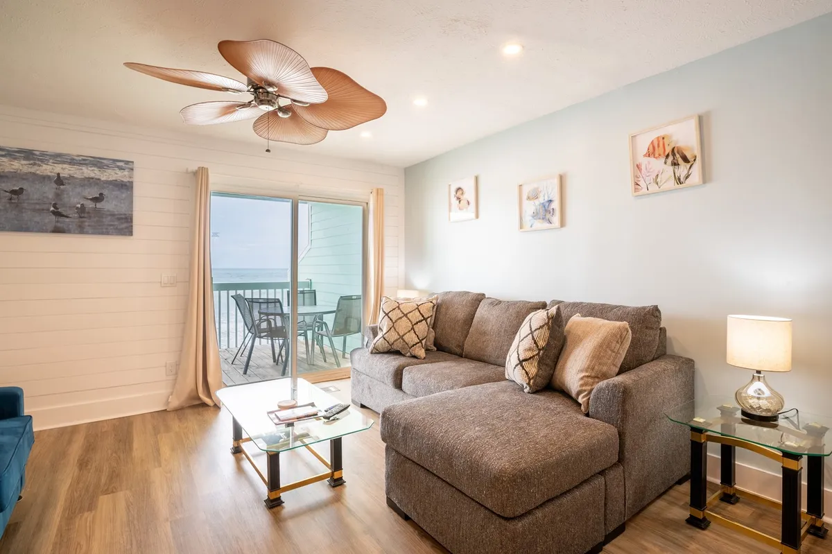 This image features a comfortable sofa and coffee table set in front of large balcony doors, with artwork on the wall and a tranquil, inviting atmosphere. The space is well-lit by natural light pouring in from the glass doors, which open directly onto a balcony with an ocean view—showcasing the seamless connection between indoors and outdoors.
