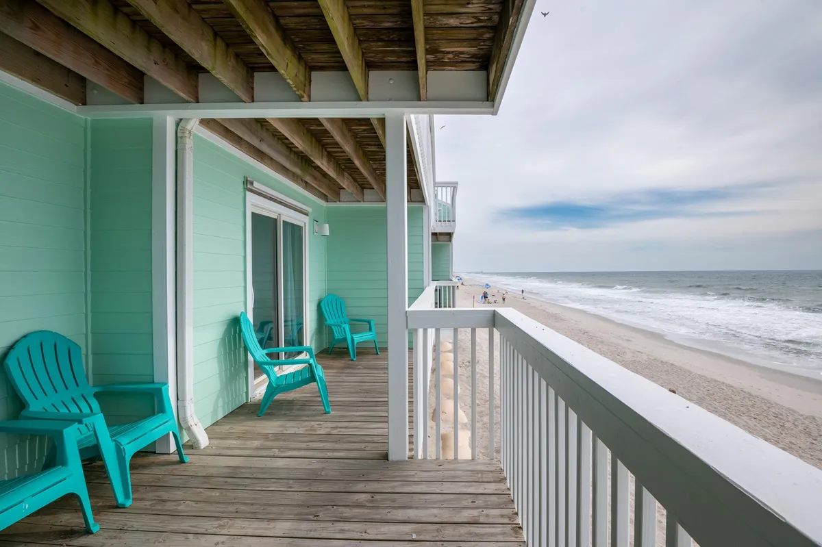 This exterior image showcases two turquoise chairs positioned on a wooden deck, overlooking a beautiful beach view. The ocean is visible in the background, and the tranquil mood is highlighted by the open sky and soft waves. The setup suggests a relaxing environment where guests can sit, read, and fully immerse themselves in the natural surroundings.