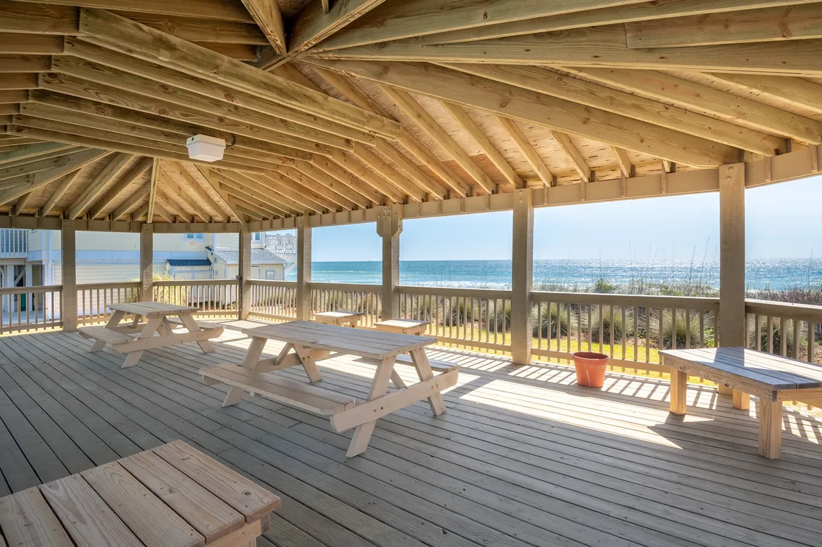 An exterior scene featuring picnic tables under wooden beams with an ocean view in the background. The area is cheerful and designed for social or family gatherings, with the beach just a few steps away. The setting encourages outdoor activities such as picnicking after a morning of hiking or exploring nearby parks.