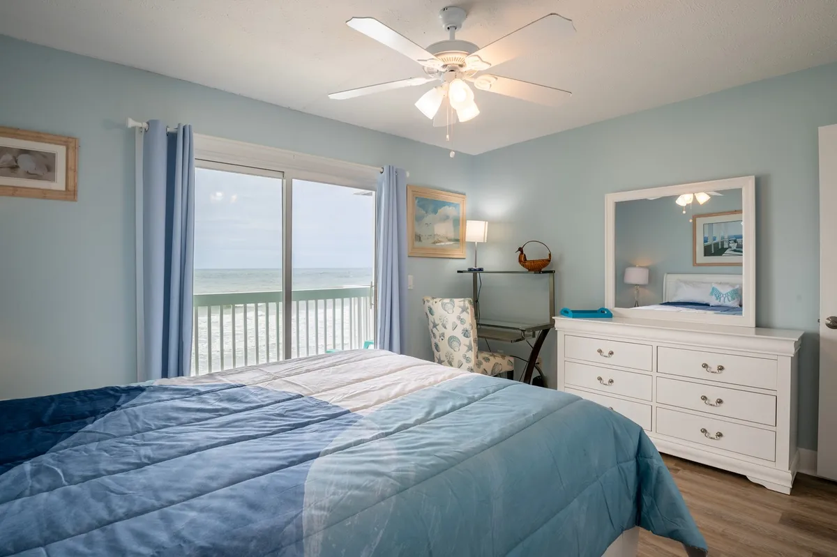 A light-filled bedroom features a comfortable queen bed with a blue blanket, a large window framing a stunning ocean view, and a dresser with mirror, creating a tranquil space perfect for unwinding. The serene palette and uncluttered layout enhance the sense of privacy and restful retreat.