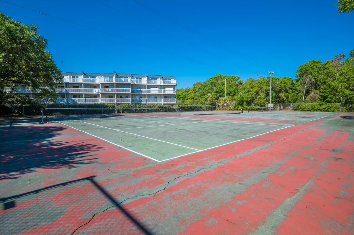 The image displays a well-maintained tennis court bordered by trees and a nearby residential building. The scene is energetic, active, and shows opportunities for outdoor exercise and fun.