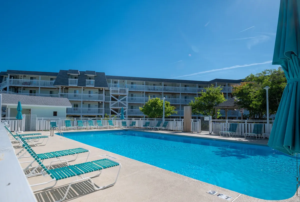 This image shows a sparkling outdoor swimming pool surrounded by lounge chairs and green trees, with the facade of surrounding resort buildings visible in the background. The atmosphere is open, leisurely, and inviting, suggesting a place to relax, socialize, and enjoy the sunshine.